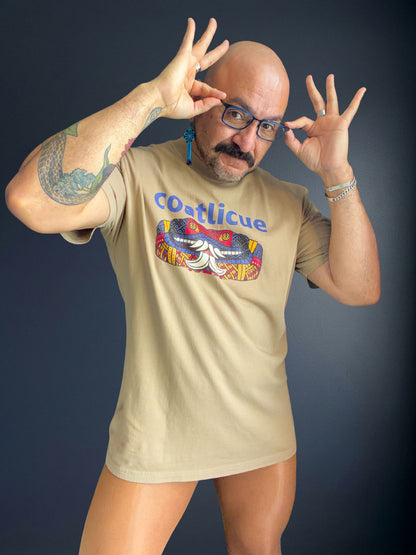 Cesar Torres wearing the faded khaki Coatlicue oversized distressed tee, both hands raised beside their glasses.